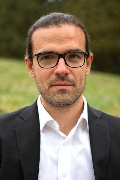 Man with dark hair pulled wearing black glasses a white collared shirt and dark blazer