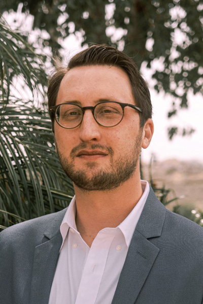 Man with dark framed glasses and short dark hair wearing a grey blazer over a shite collared shirt