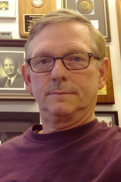 Man with glasses wearing a burgundy sweater with a background of framed plaques.