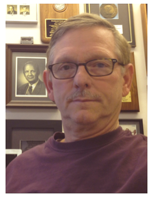 Man with glasses wearing a burgundy sweater with a background of framed plaques.